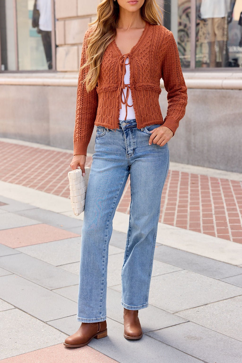 Woman wearing the Reese Tie Front Sweater - Terracotta, white top, blue jeans, brown boots, and holding a white woven clutch on sidewalk.