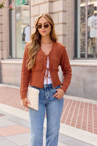 Woman in sunglasses wears the Reese Tie Front Sweater - Terracotta with jeans, holding a woven clutch on a city sidewalk.
