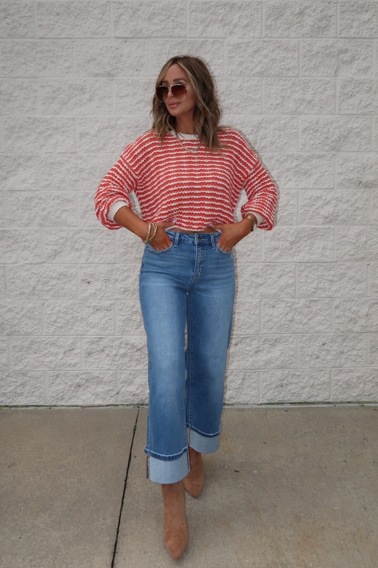Woman wearing the Reina Coral Textured Stripe Sweater, wide-leg cuffed jeans, brown heels, and sunglasses by a brick wall.