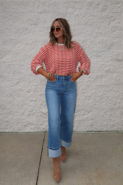 Woman wearing the Reina Coral Textured Stripe Sweater, wide-leg cuffed jeans, brown heels, and sunglasses by a brick wall.