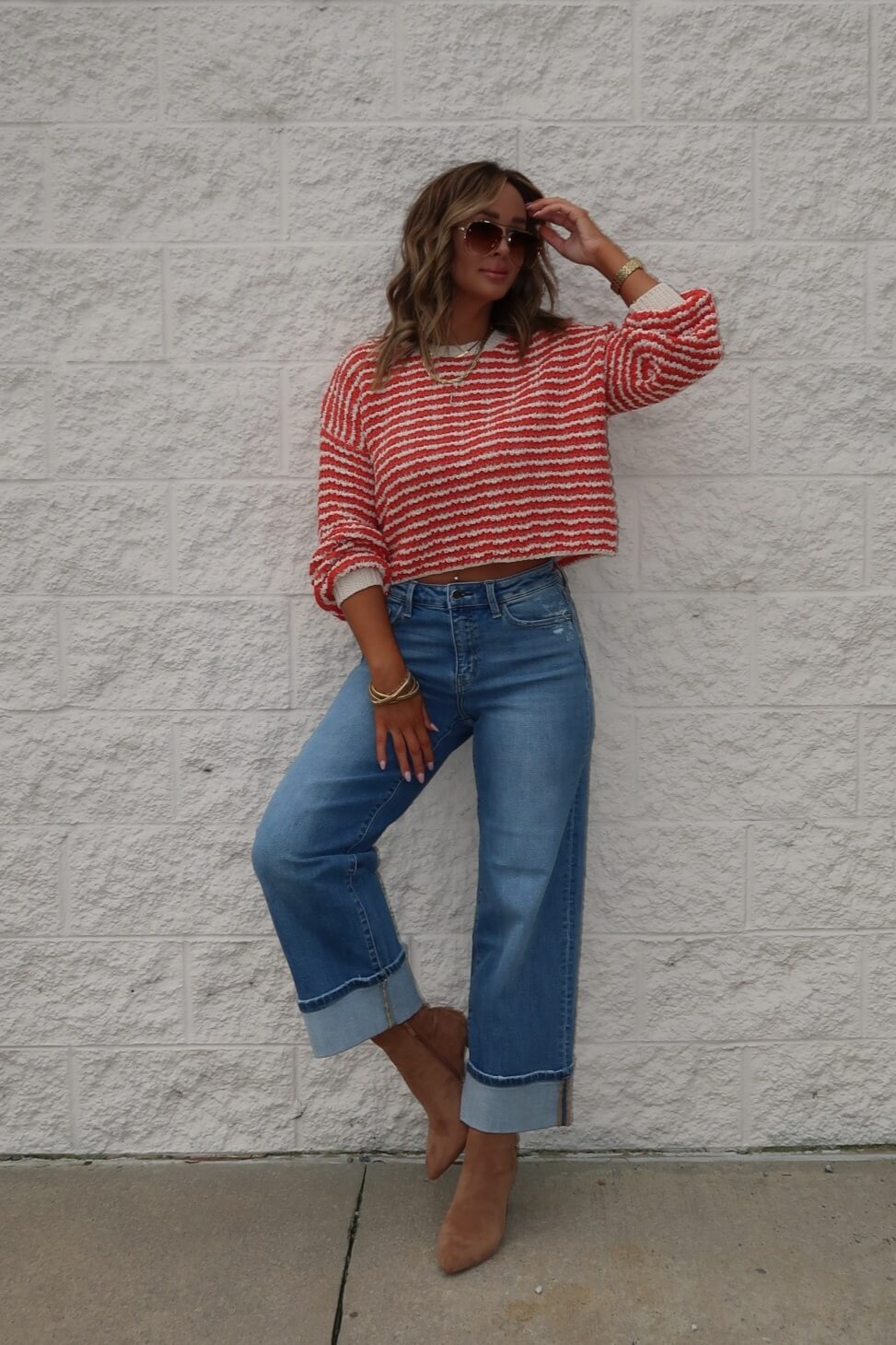 A woman in sunglasses wears the Reina Coral Textured Stripe Sweater, wide-leg cuffed jeans, and tan heeled boots by a white brick wall.
