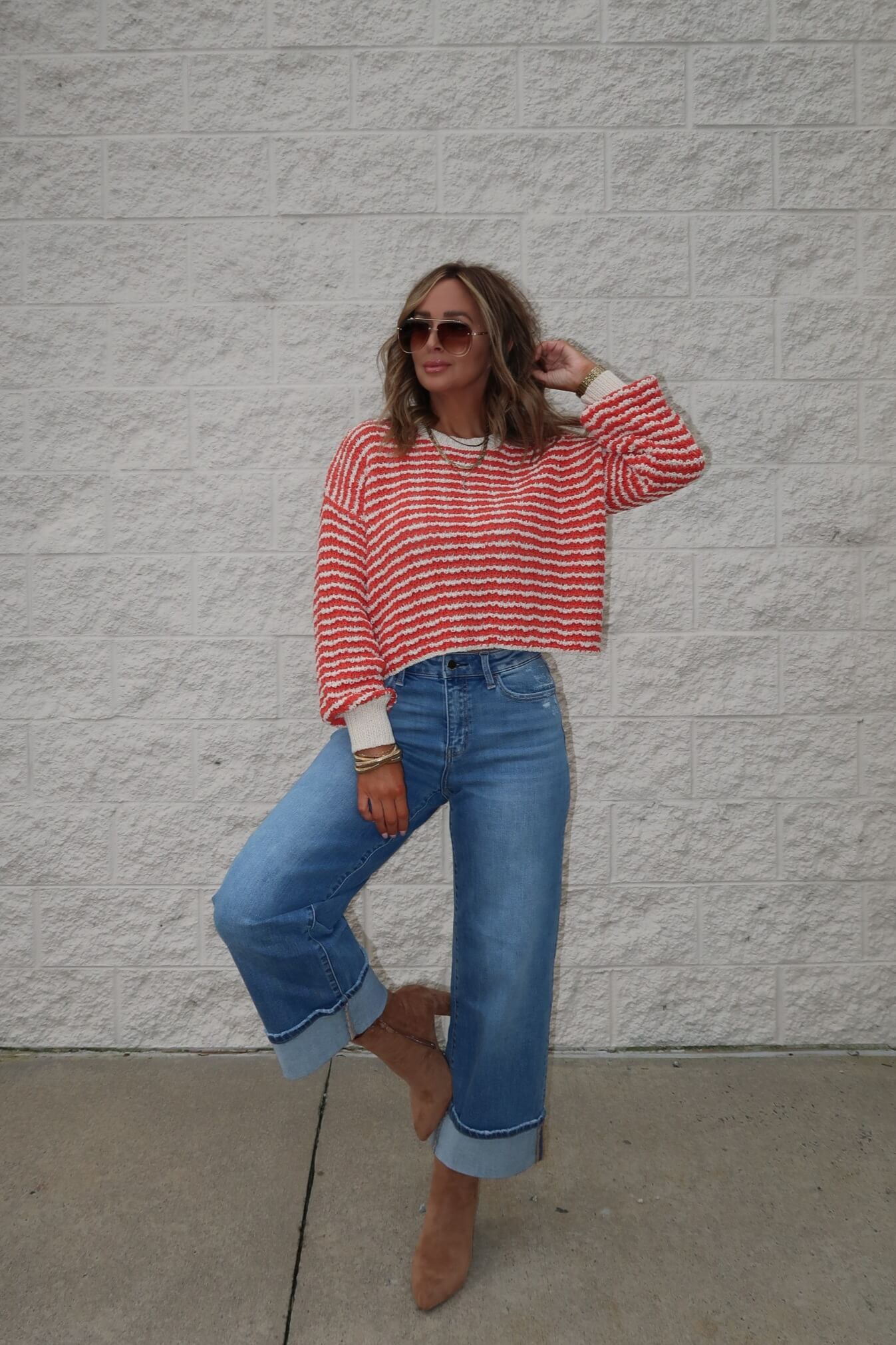 Woman poses by a white brick wall in sunglasses, wide-leg jeans, tan boots, and the Reina Coral Textured Stripe Sweater—ideal for fall.
