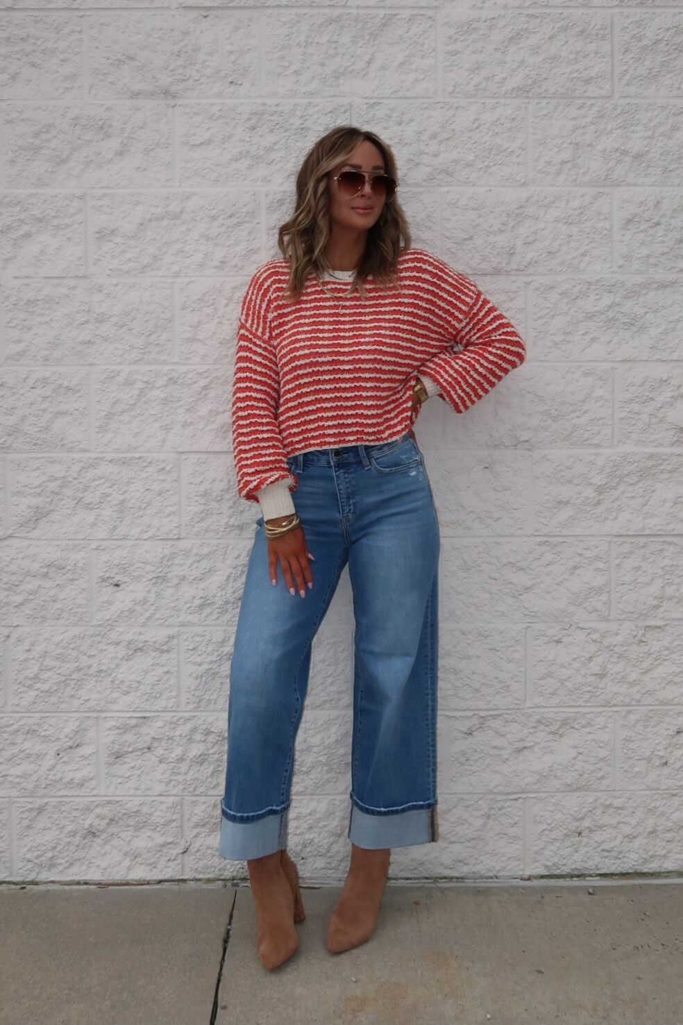 Woman wears the Reina Coral Textured Stripe Sweater, wide-leg jeans, and tan heels, posing by a light brick wall with hand on hip.