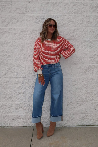 Woman wears the Reina Coral Textured Stripe Sweater, wide-leg jeans, and tan heels, posing by a light brick wall with hand on hip.