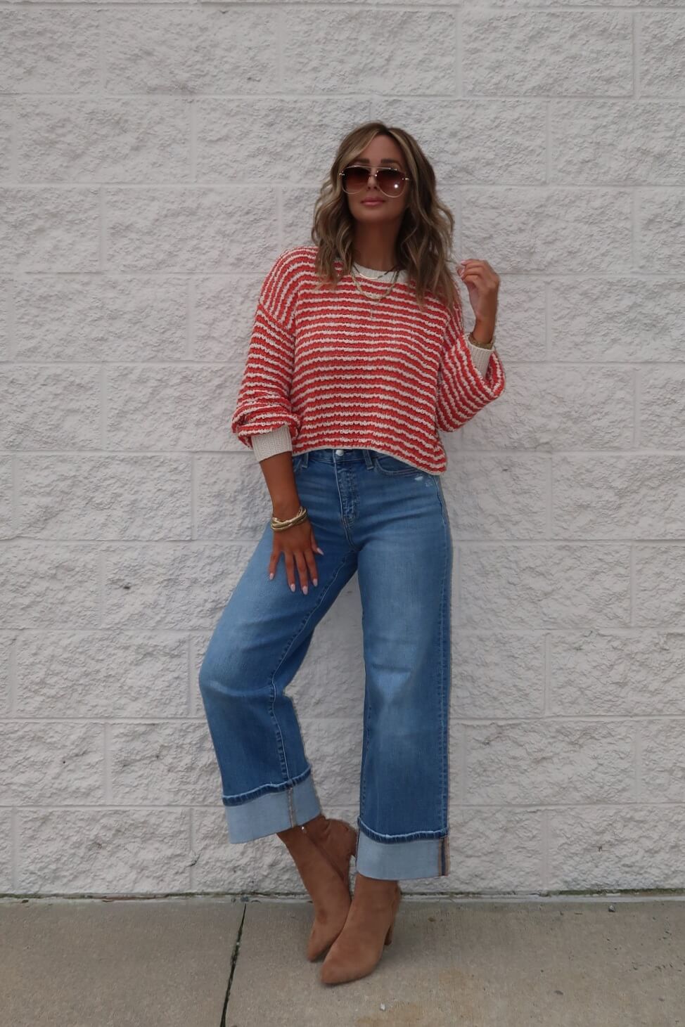 Woman in a Reina Coral Textured Stripe Sweater, wide-leg cuffed jeans, brown boots, and sunglasses stands by a light brick wall.