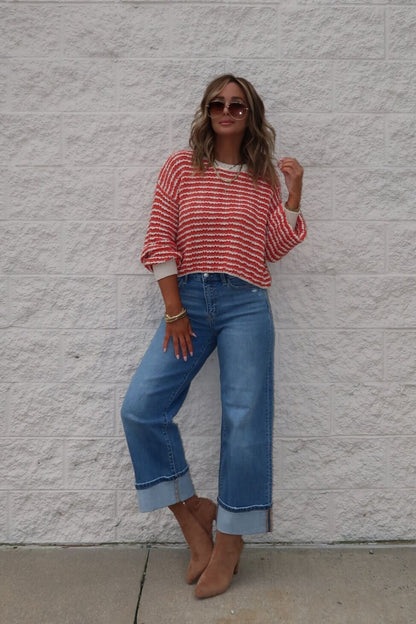 Woman in a Reina Coral Textured Stripe Sweater, wide-leg cuffed jeans, brown boots, and sunglasses stands by a light brick wall.