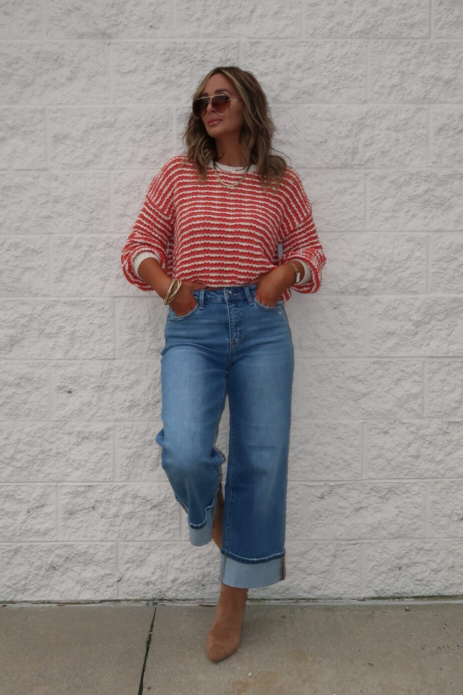 Woman in sunglasses wears the Reina Coral Textured Stripe Sweater with wide-leg jeans and tan boots by a textured white wall.