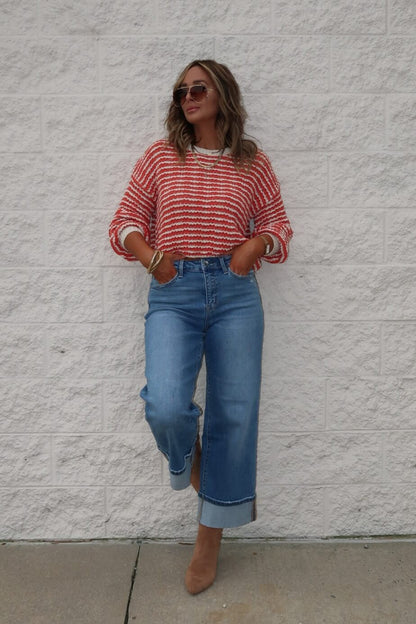 Woman in sunglasses wears the Reina Coral Textured Stripe Sweater with wide-leg jeans and tan boots by a textured white wall.