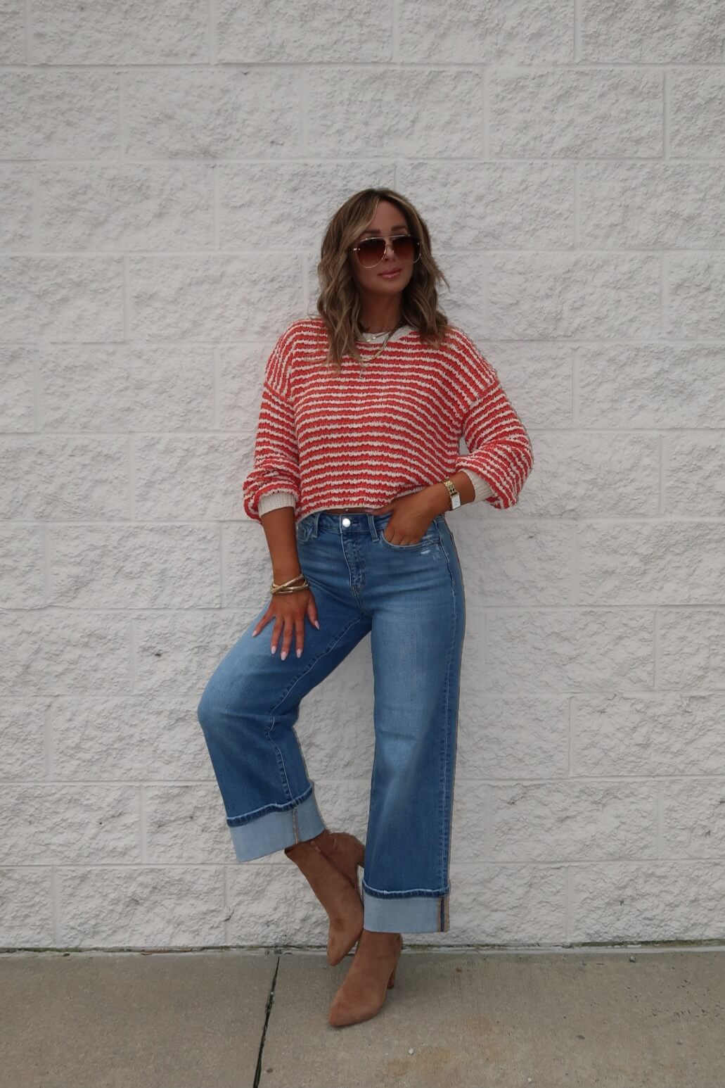 A woman in sunglasses wears the Reina Coral Textured Stripe Sweater with cuffed blue jeans and tan ankle boots.