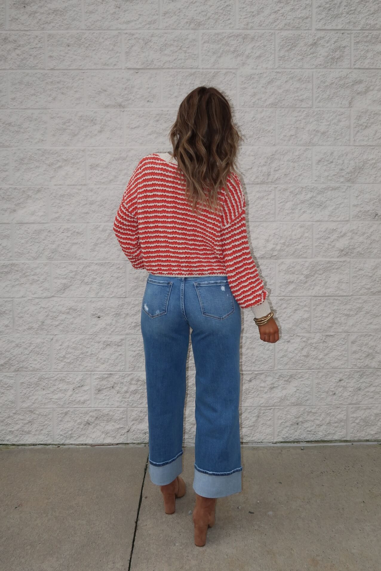 A woman with wavy hair faces a white brick wall, wearing the Reina Coral Textured Stripe Sweater and cuffed blue jeans.