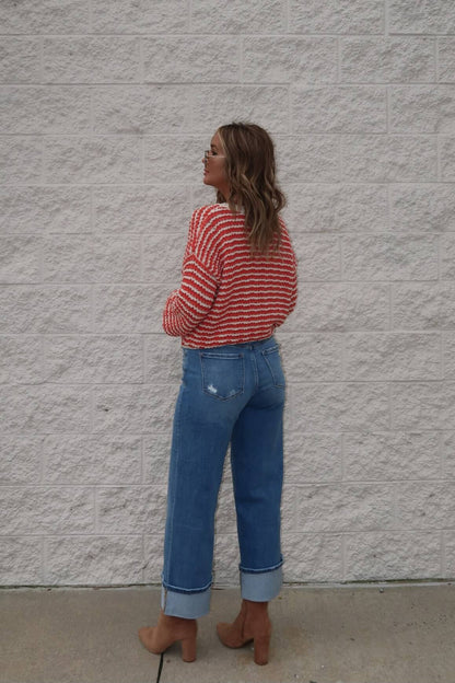A woman in a Reina Coral Textured Stripe Sweater, blue rolled-up jeans, and brown shoes faces a textured white brick wall.