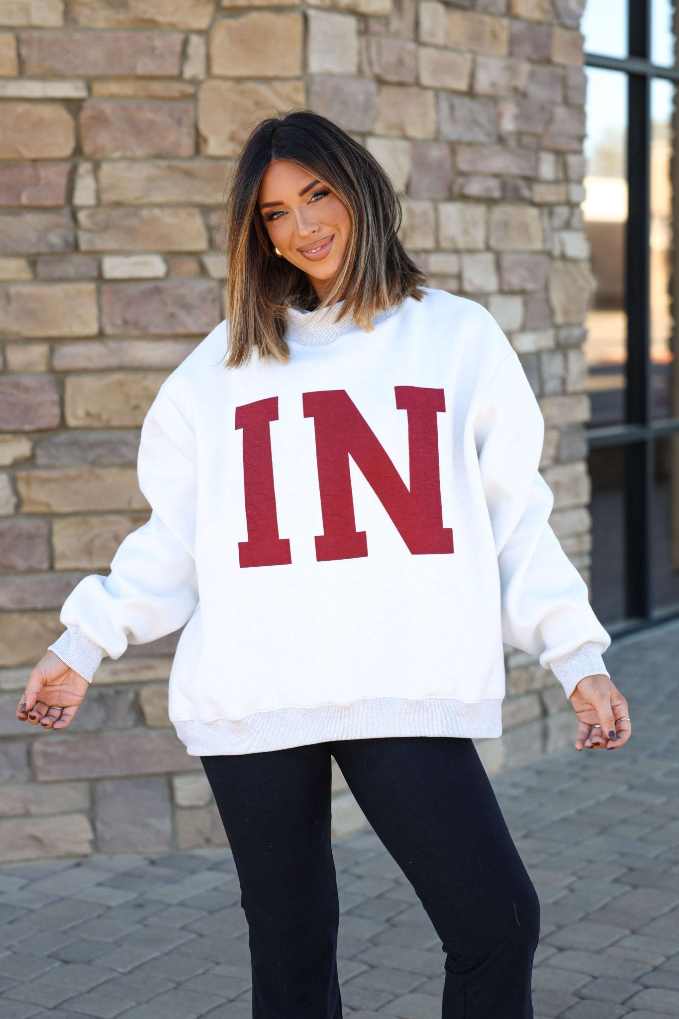 Woman smiles in a Reversible Indiana Pullover Sweatshirt with bold red IN letters, radiating Hoosier pride by a brick wall.