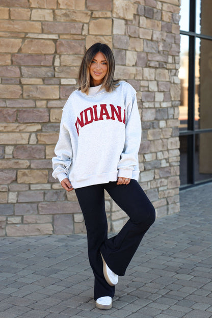 Woman smiles outdoors in a gray Reversible Indiana Pullover Sweatshirt, black pants, and white shoes by a stone wall.