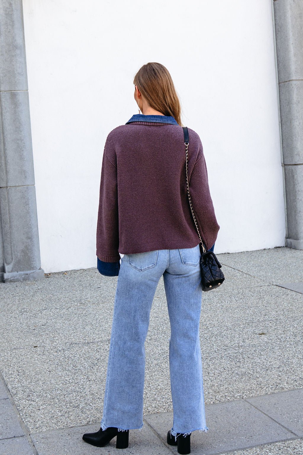 A woman in a Ribbed Denim Contrast Sweater - Mocha, light blue jeans, and black shoes stands facing a white wall outdoors.