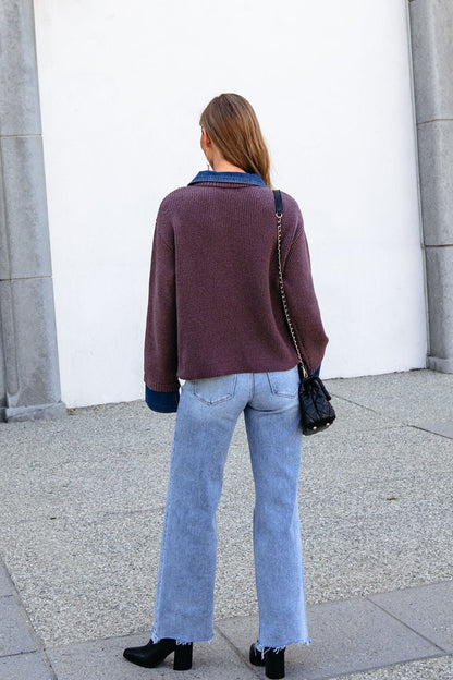 A woman in a Ribbed Denim Contrast Sweater - Mocha, light blue jeans, and black shoes stands facing a white wall outdoors.