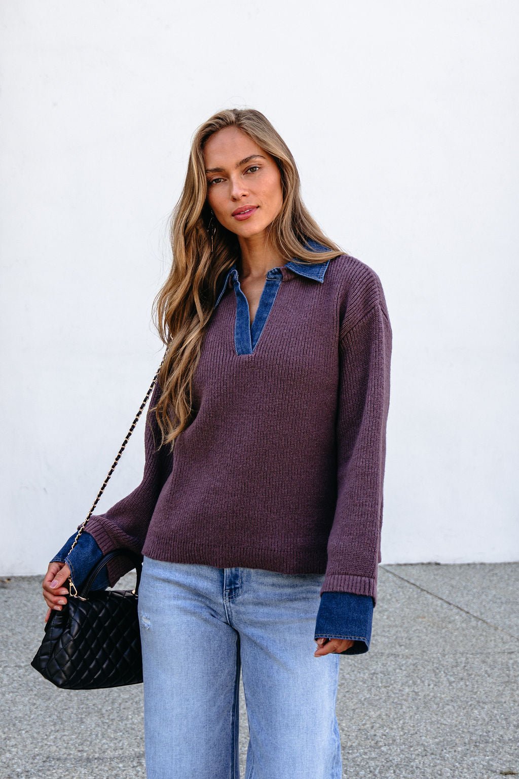 Woman with long hair in a Ribbed Denim Contrast Sweater - Mocha, light jeans, and a black purse stands against a white wall.
