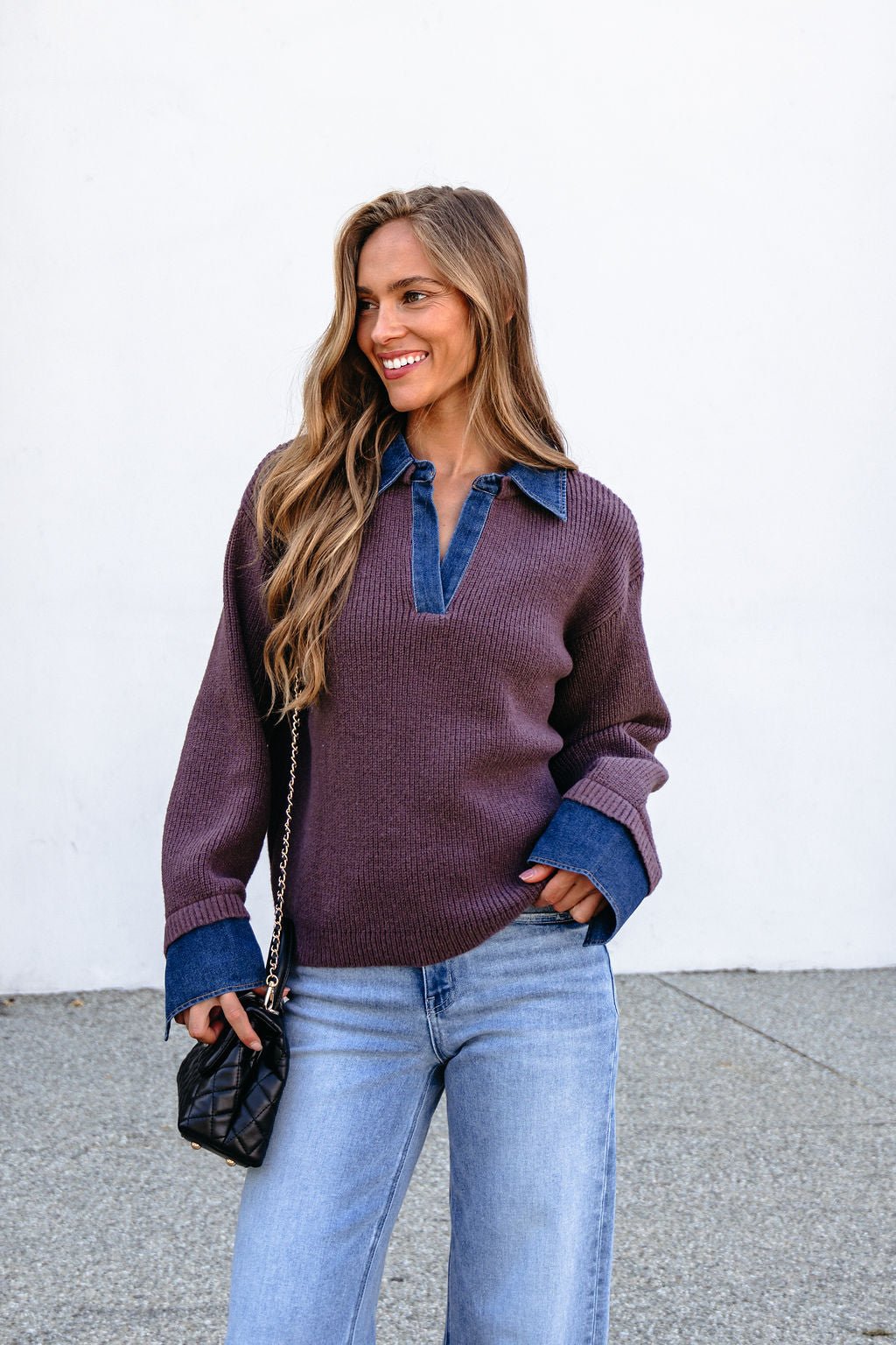 Woman smiles in a Ribbed Denim Contrast Sweater - Mocha over a denim shirt, light blue jeans, with a small black purse outdoors.