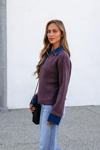 Woman with long hair in a Ribbed Denim Contrast Sweater - Mocha and jeans stands outdoors, holding a black purse and looking at the camera.