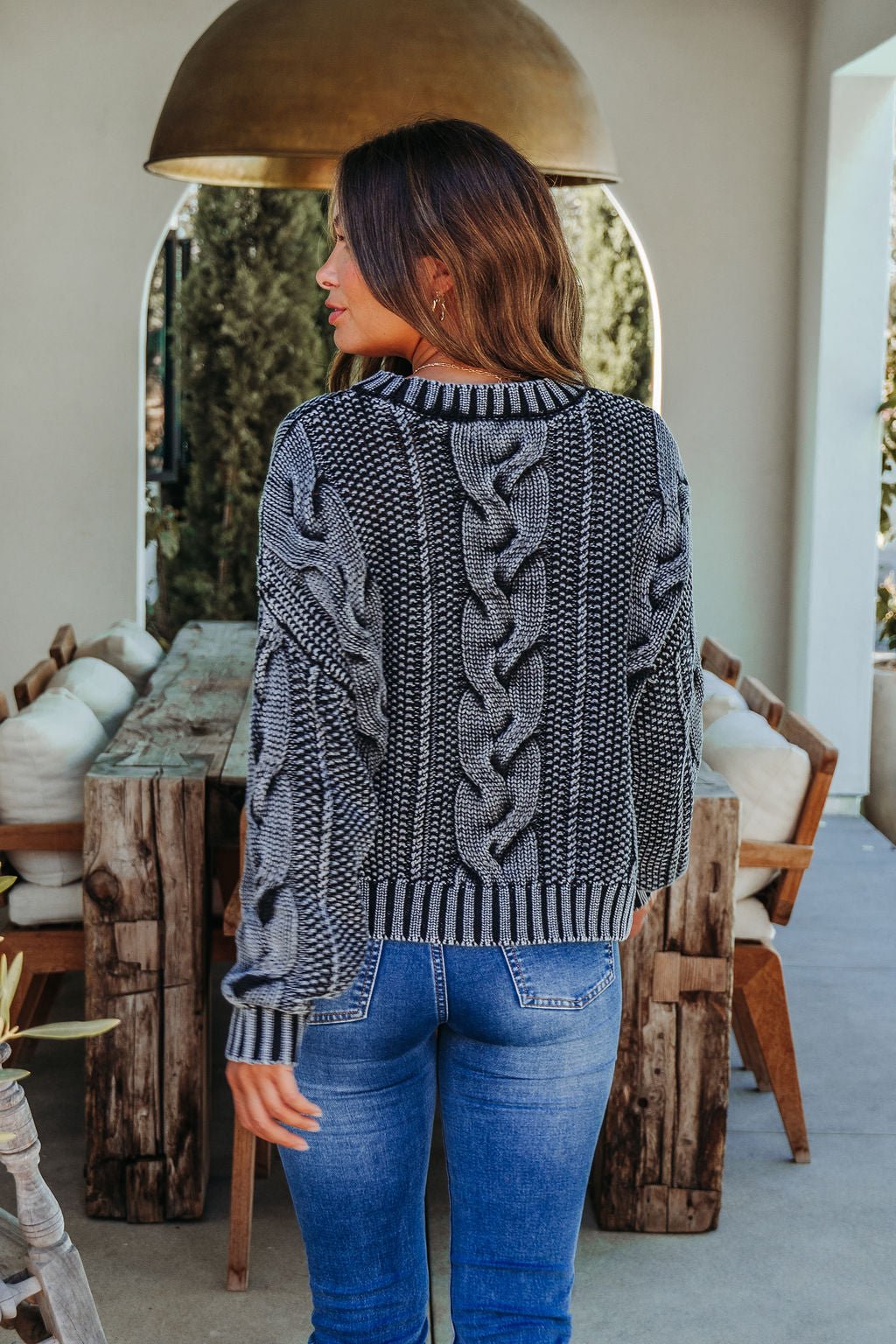 A woman with wavy brown hair stands indoors, back to the camera, wearing the Riley Charcoal Cable Knit Sweater and blue jeans.