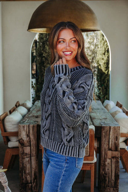 Woman wearing the Riley Charcoal Cable Knit Sweater and jeans stands by a rustic wooden dining table, smiling to the side.