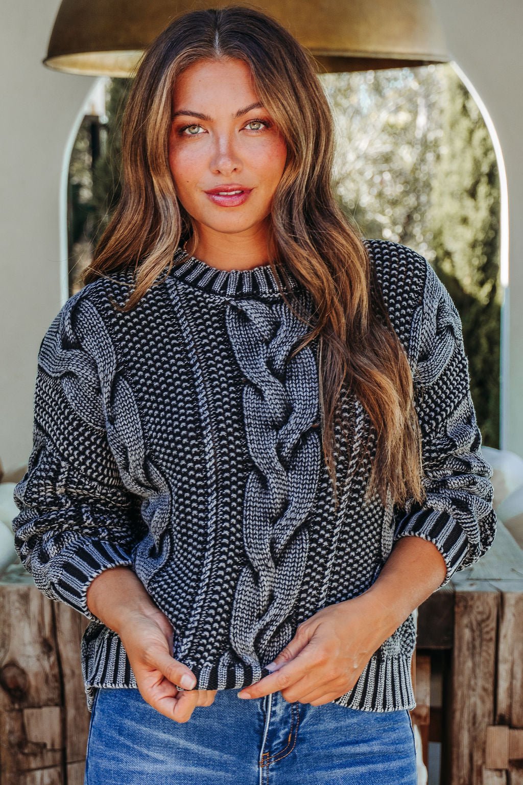 Woman with long brown hair wearing the Riley Charcoal Cable Knit Sweater and blue jeans, standing outdoors in natural light.