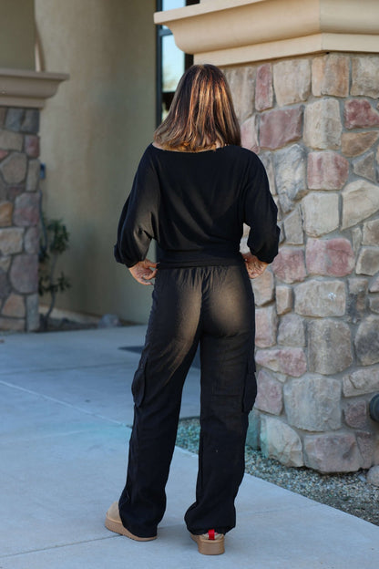 A woman with shoulder-length hair stands outside near a stone wall wearing Risen Black Wide Leg Cargo Pants.
