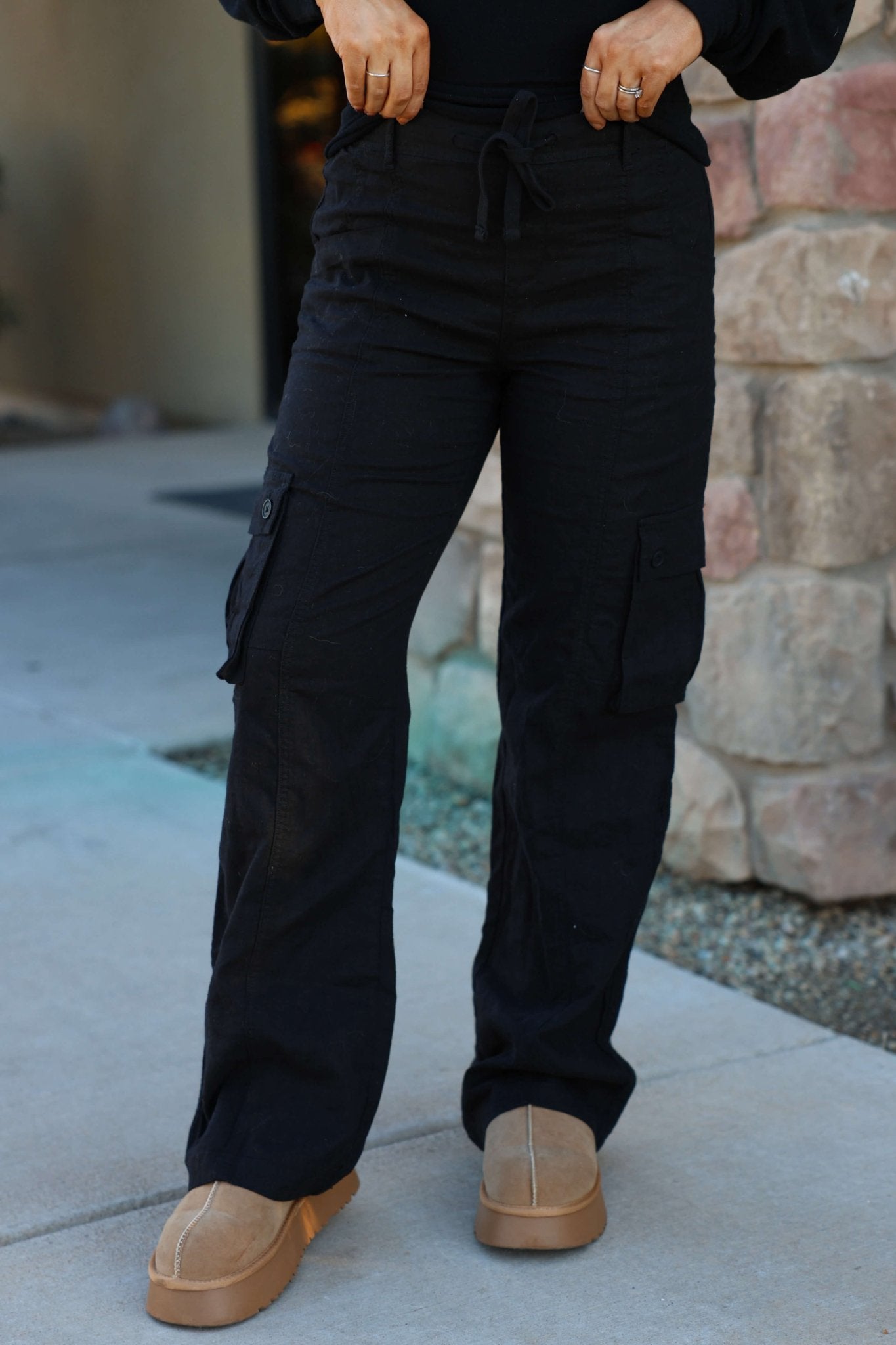Person standing outdoors in Risen Black Wide Leg Cargo Pants with side pockets; tan shoes, hands on waistband, stone wall behind.