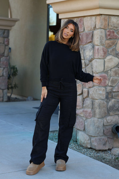 A woman wears Risen Black Wide Leg Cargo Pants with side pockets, standing by a stone wall and smiling casually outdoors.