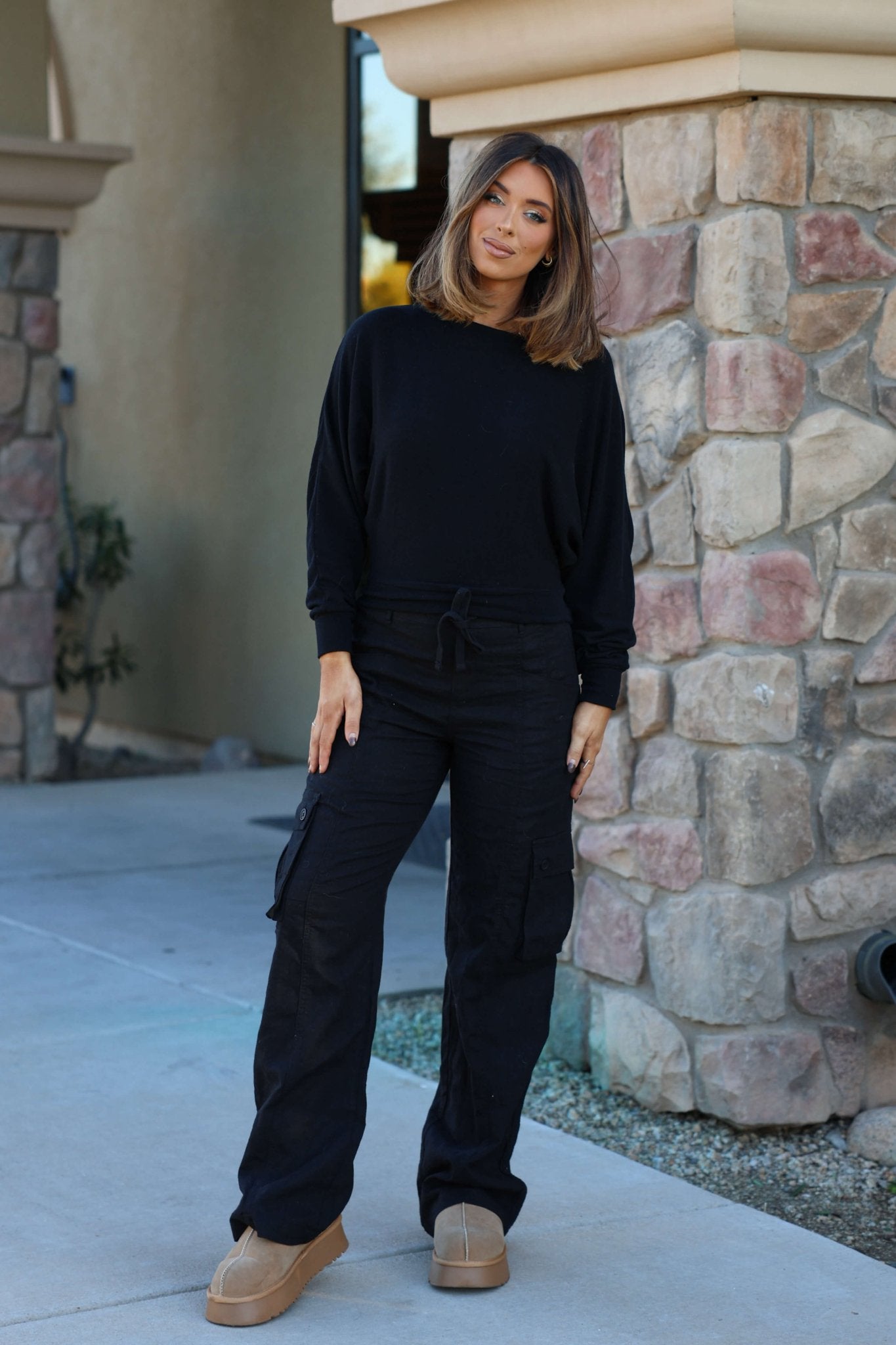 A woman stands outside by a stone wall wearing Risen Black Wide Leg Cargo Pants with side cargo pockets, looking at the camera.
