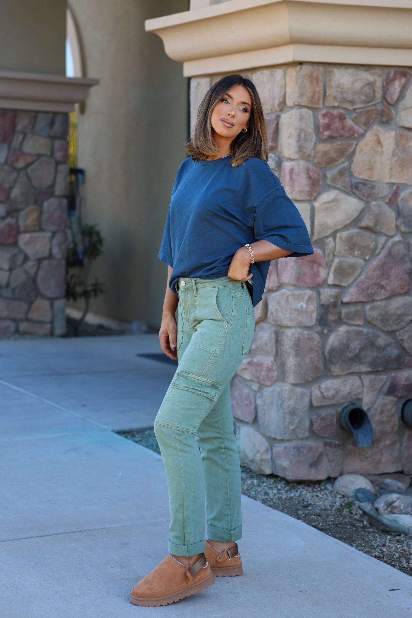 Woman by a stone wall wears Risen Olive Straight Leg Cargo Pants, a blue top, and tan shoes, looking at the camera.
