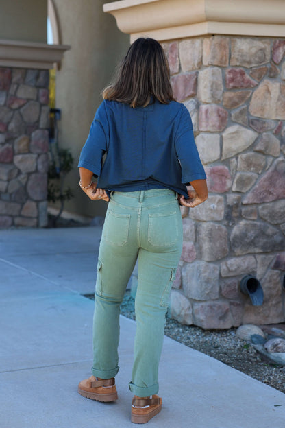 A woman stands outside on a sidewalk, wearing Risen Olive Straight Leg Cargo Pants, a blue top, and brown shoes.