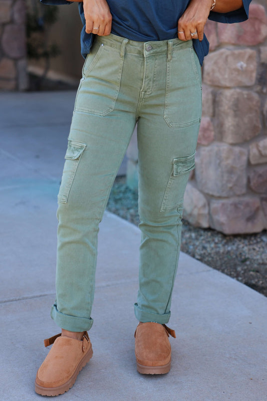 Wearing Risen Olive Straight Leg Cargo Pants and tan shoes, a person stands on a sidewalk with hands on waistband, stone wall behind.