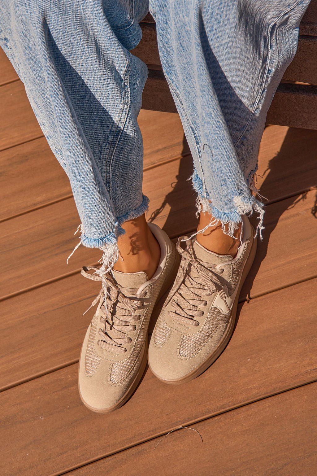 Person wearing Riviere Taupe Raffia Sneakers and frayed light blue jeans, sitting on a wooden surface in sunlight.
