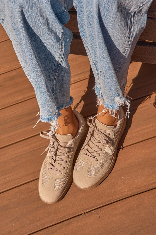 Person wearing Riviere Taupe Raffia Sneakers and frayed light blue jeans, sitting on a wooden surface in sunlight.