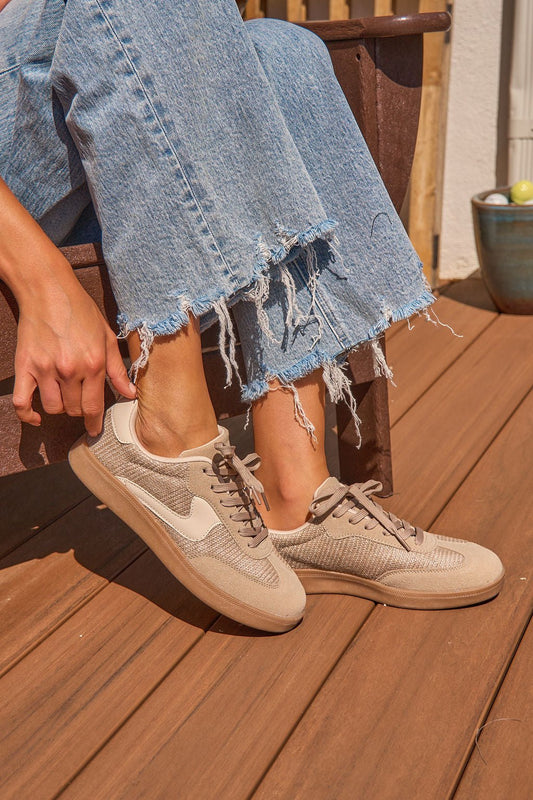 Person wearing frayed hem jeans and Riviere Taupe Raffia Sneakers, sitting on a wooden deck, adjusting one shoe.