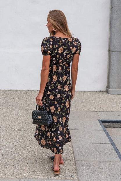 Woman with long hair walks away in the Rosalie Floral Midi Dress - Black, holding a black purse on a stone path.