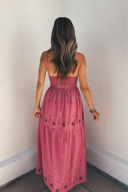 Woman with long wavy hair in the Rosalie Pink Embroidered Halter Midi Dress stands facing a plain wall.