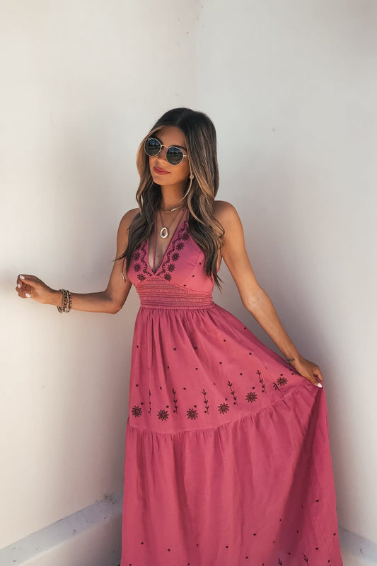 Woman in sunglasses wears the Rosalie Pink Embroidered Halter Midi Dress, standing by a white wall and holding her dress.