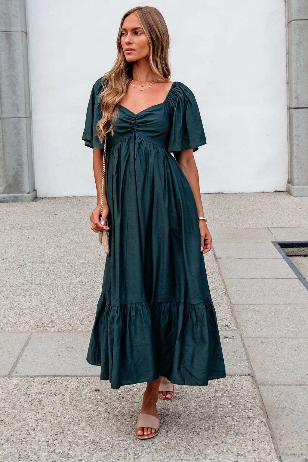 Woman in the Rosalind Sweetheart Smocked Midi Dress—Green stands outdoors on a stone walkway, sandals and crossbody bag completing her look.