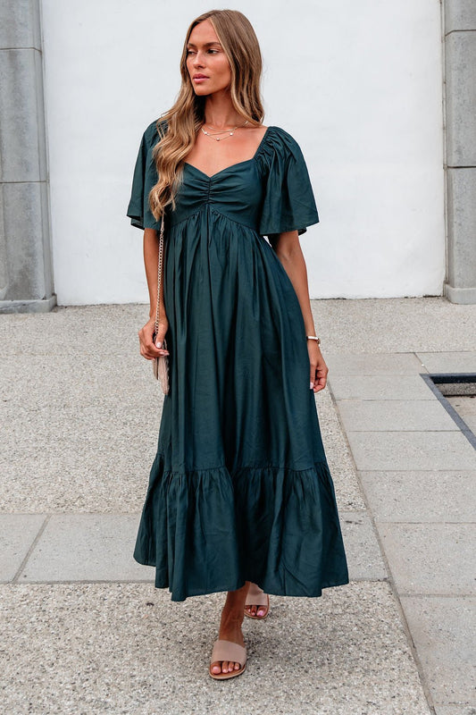 Woman in the Rosalind Sweetheart Smocked Midi Dress—Green stands outdoors on a stone walkway, sandals and crossbody bag completing her look.