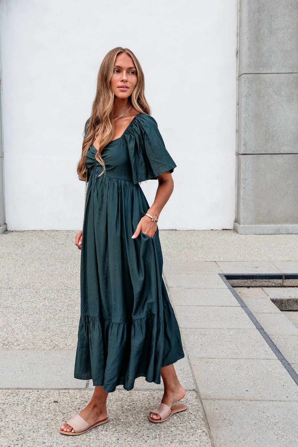 Woman in the Rosalind Sweetheart Smocked Midi Dress—Green, stands outdoors on a concrete path, styled with sandals.