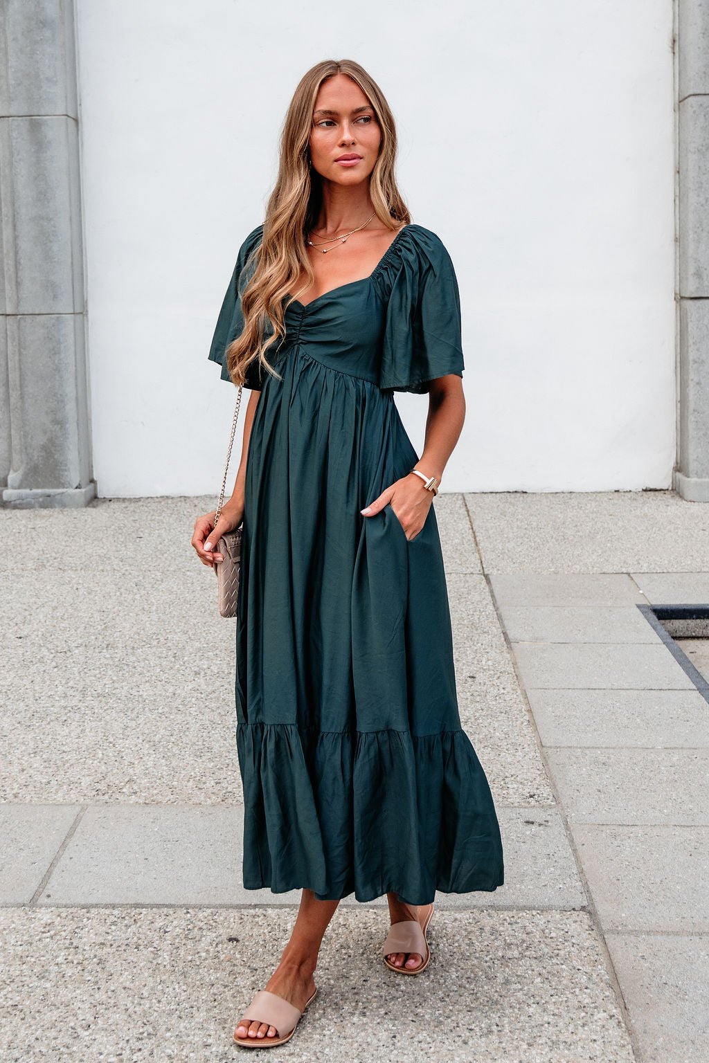 Woman in the Rosalind Sweetheart Smocked Midi Dress—Green, with sandals and a purse, stands outdoors on a stone path.
