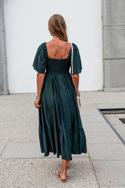 Woman in Rosalind Sweetheart Smocked Midi Dress — Green walks away on a paved path, carrying a light purse and wearing sandals.