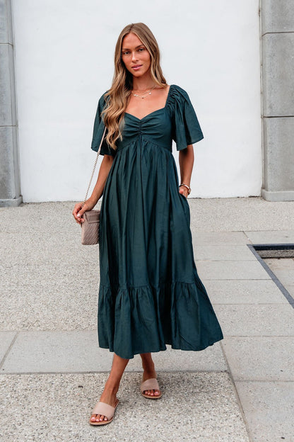 Woman in the Rosalind Sweetheart Smocked Midi Dress — Green, sandals, and a beige purse stands on a concrete walkway.