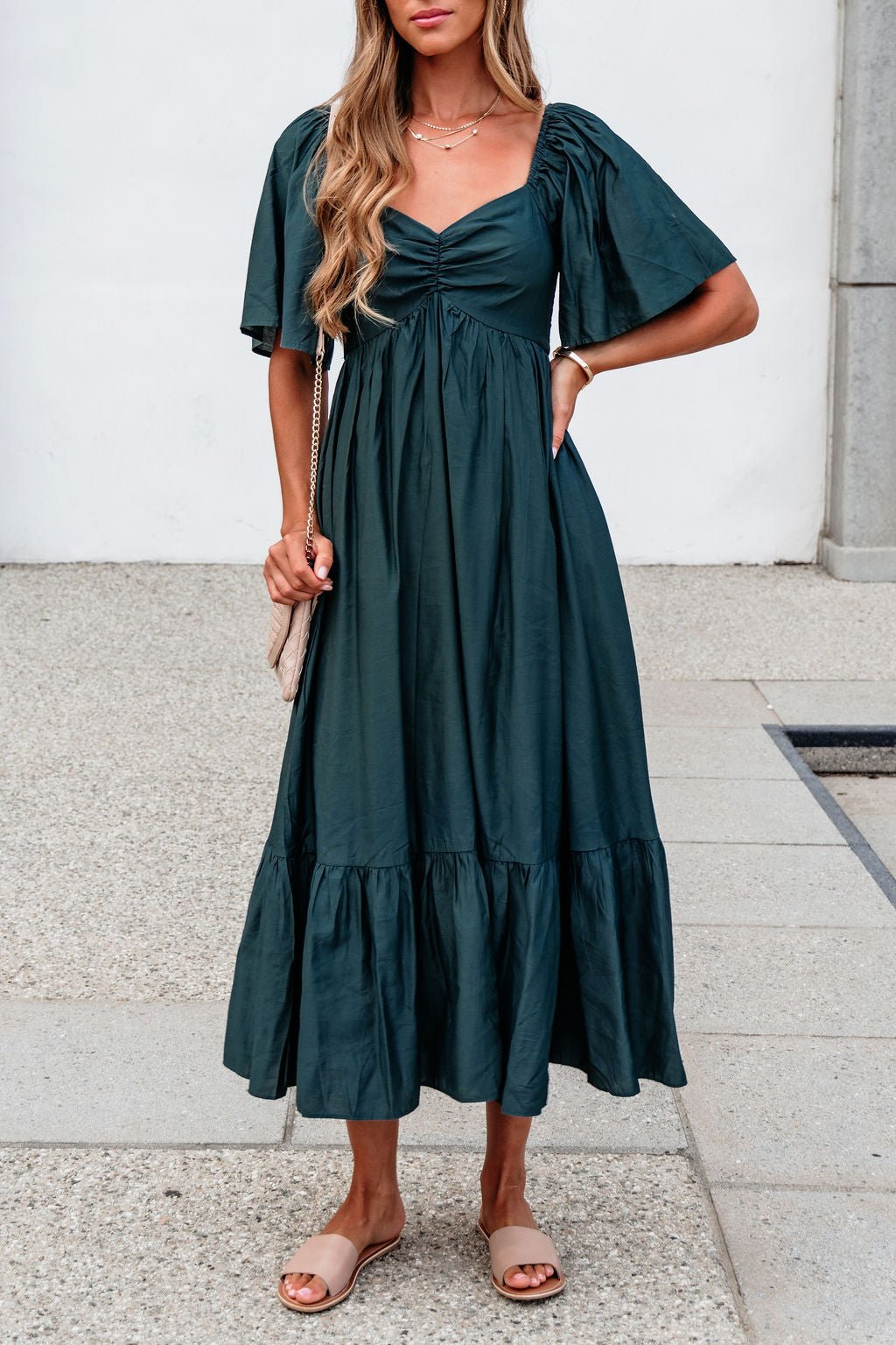 Woman in a Rosalind Sweetheart Smocked Midi Dress — Green, beige sandals, and a small beige purse on a stone walkway.