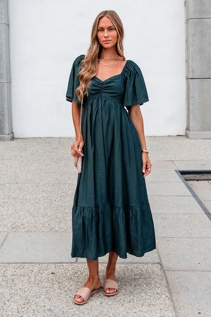 Woman in the Rosalind Sweetheart Smocked Midi Dress — Green stands on a stone walkway, pairing it with sandals and a light purse.