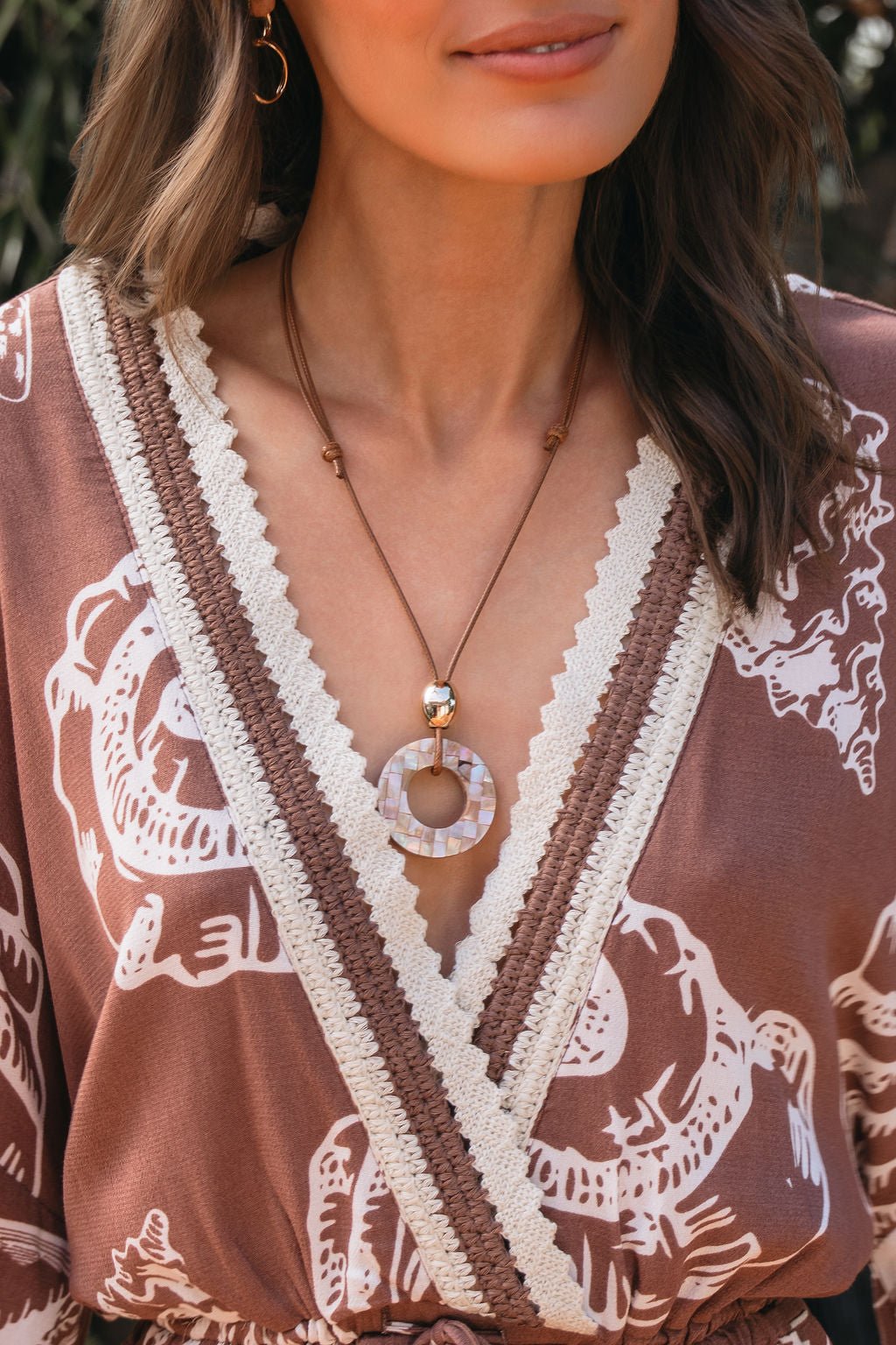 A woman wears a patterned brown dress with a deep V-neck and the Rosé Coast Mosaic Pendant Necklace.
