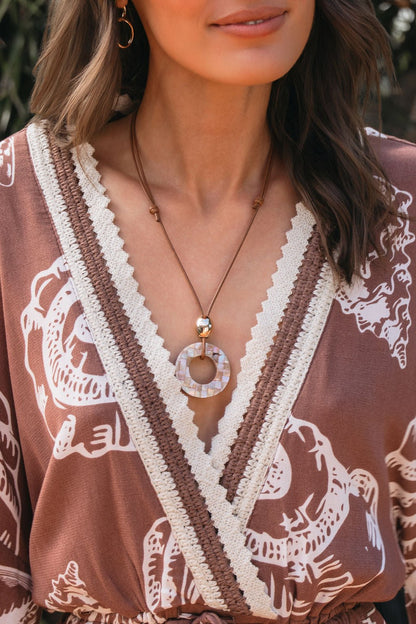 A woman wears a patterned brown dress with a deep V-neck and the Rosé Coast Mosaic Pendant Necklace.