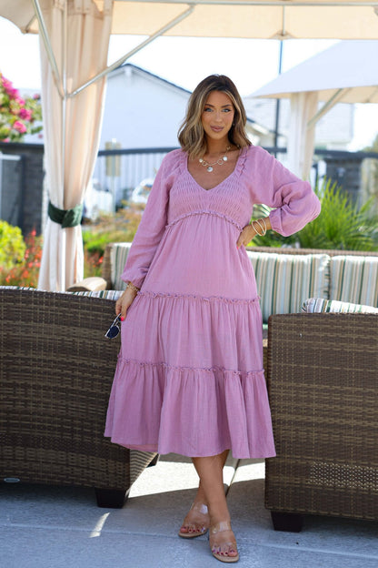A woman in the Rosy Pink Smock Tiered Midi Dress stands outdoors by wicker furniture, holding sunglasses near a canopy and greenery.