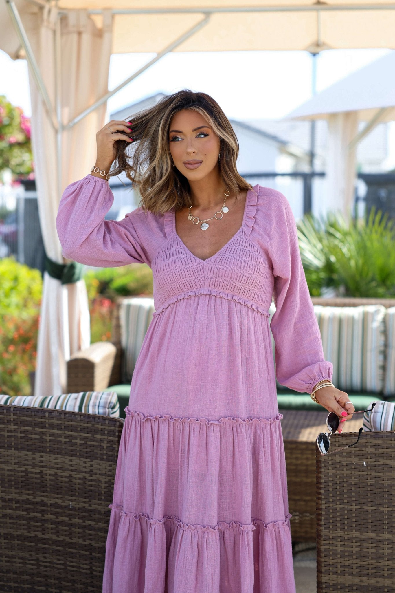 Woman with wavy hair poses outdoors under a canopy in the Rosy Pink Smock Tiered Midi Dress, holding her hair and sunglasses.